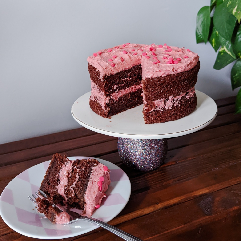 A very cute chocolate layer cake with pink frosting and pink sprinkles and a slice removed and partially eaten.